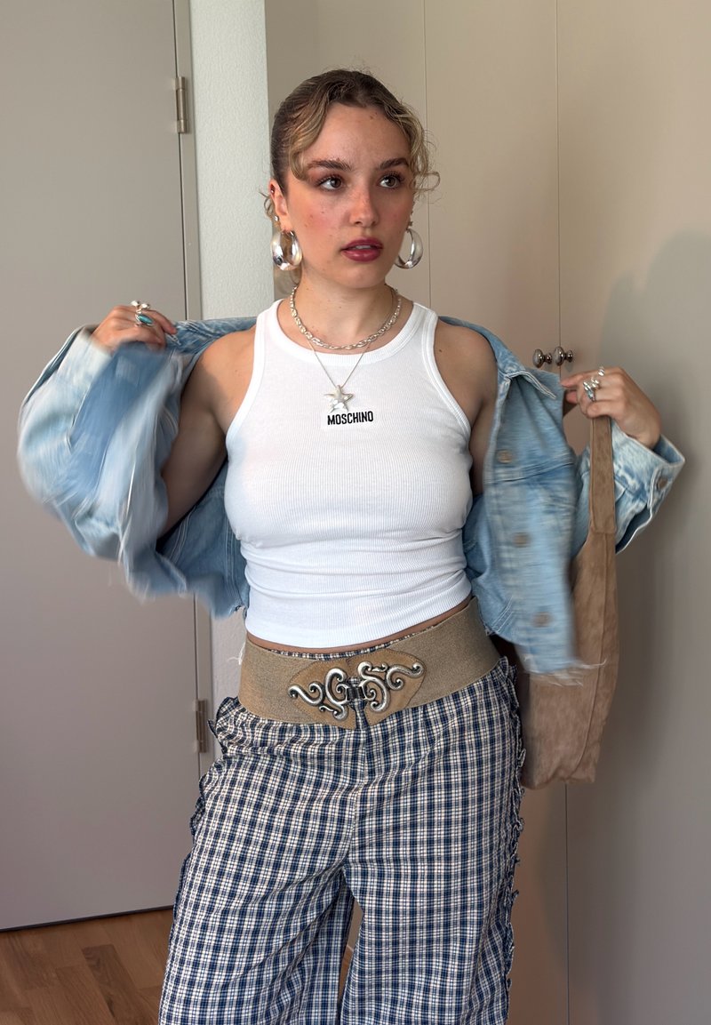 Jeune femme ajustant une veste en jean bleu clair, portant un top blanc sans manches avec un collier Moschino, un pantalon à carreaux et de grandes boucles d'oreilles créoles.