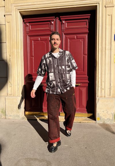 Camisa de manga corta con un patrón en blanco y negro, pantalones de pierna ancha rojos oscuros y zapatos negros. De pie frente a una puerta decorativa roja.