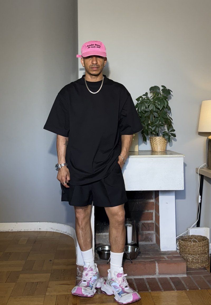 Homme portant un ensemble t-shirt noir oversize et short, casquette rose, chaussettes blanches et baskets épaisses roses et blanches, debout à l'intérieur près d'une cheminée.