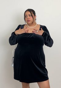 Rochie din catifea neagră, cu decolteu pătrat, mâneci bufante și siluetă lejeră. Accentuată cu o bandă subțire aurie și bijuterii din perle.