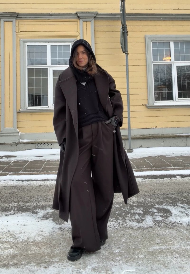 Donna in piedi all'aperto su una strada innevata, con indosso un lungo cappotto scuro, pantaloni larghi, felpa nera con cappuccio, guanti e stivali invernali, con uno sfondo di edificio giallo.