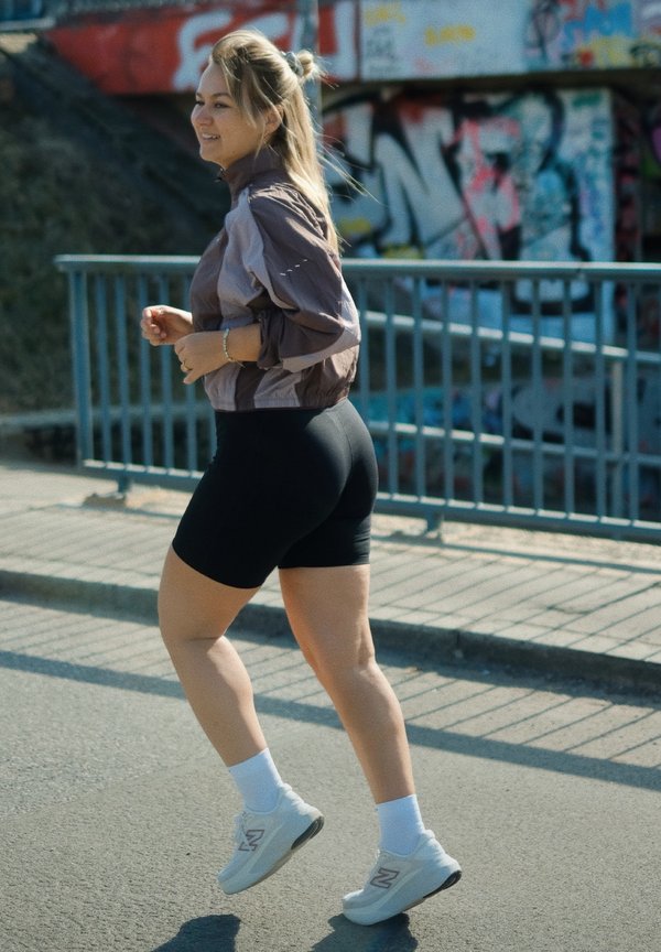 Giovane donna che fa jogging all'aperto, indossando una giacca leggera, pantaloncini neri, calzini bianchi e scarpe da ginnastica vicino a un muro coperto di graffiti.