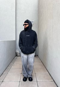 Μαύρη, φαρδιά κουκούλα με μπροστινή τσέπη και λογότυπο, σε συνδυασμό με ανοιχτό γκρι παντελόνι σε φαρδιά γραμμή και μαύρα παντελόνια χωρίς λουριά, στέκεται σε έναν στενό διάδρομο.