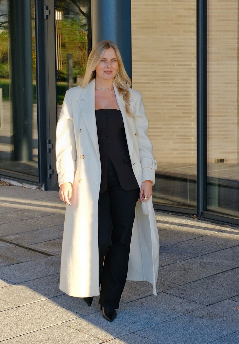 Long cream overcoat, fitted black top with buttons, and black trousers. Smooth textures, sharp collar, and pointed shoes. Outdoor setting.