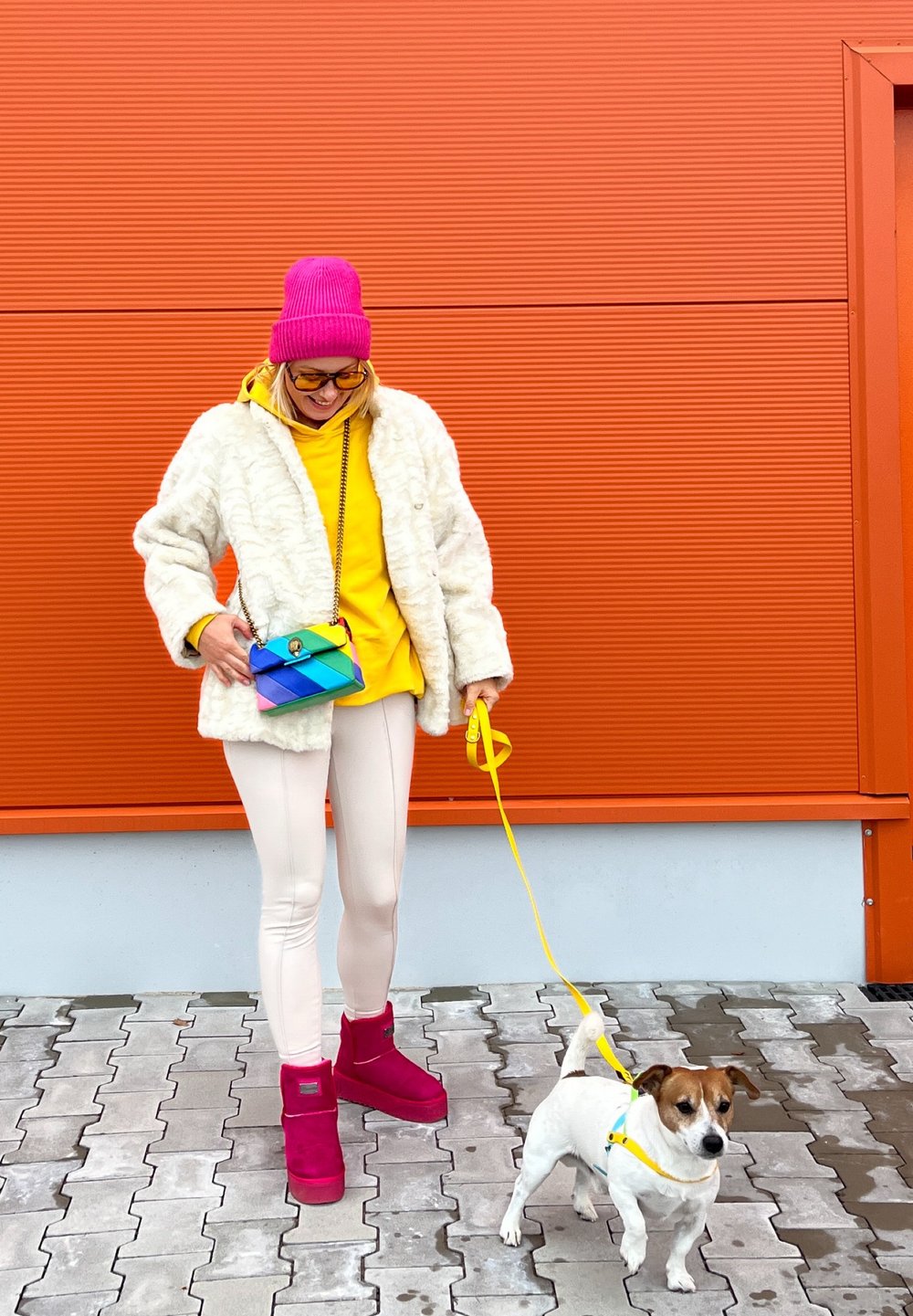 Biely kožúšok z umelého kožucha, žltá mikina s kapucňou, ružová čiapka, ružové členkové topánky a viacfarebná crossbody taška. Na vodítku je malý biely pes.
