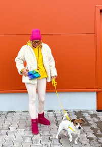 Biely kožúšok z umelého kožucha, žltá mikina s kapucňou, ružová čiapka, ružové členkové topánky a viacfarebná crossbody taška. Na vodítku je malý biely pes.