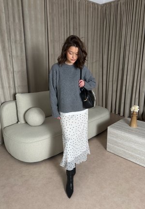 Mujer con suéter gris, falda blanca con lunares, botas negras, sosteniendo un bolso negro, de pie en una habitación moderna con sofá curvado y cortinas beige.