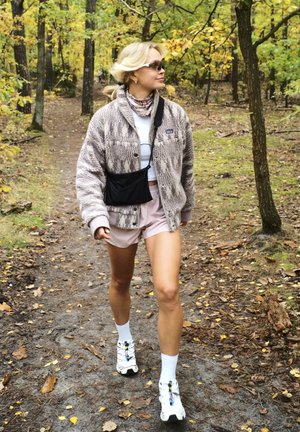 Chaqueta de forro polar en beige con franjas texturizadas, top blanco, pantalones cortos rosas y bolsa cruzada negra. Lleva calcetines blancos y zapatillas deportivas coloridas.