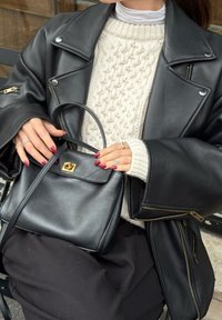 Musta nahkatakki hopeanvärisillä yksityiskohdilla, yhdistettynä kermaiseen neulepuseroon; kädessä pieni musta nahkainen käsilaukku, jossa kultainen lukko.