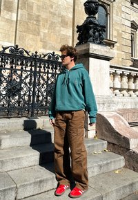 Zelena dukserica s kapuljačom, smešene cargo hlače i crvene patike sa belim prugama. Stoji na kamenim stepenicama pored dekorativnih gvozdenih vrata.
