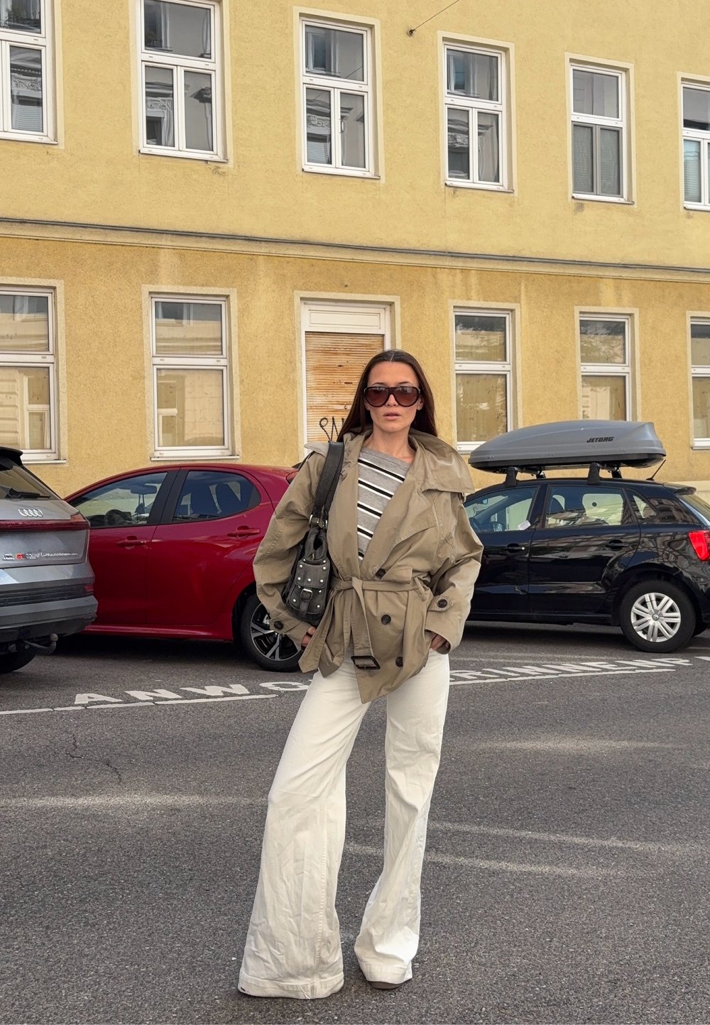 Napszemüveget viselő nő bézs kabátban, csíkos pólóban és fehér, széles nadrágban áll egy utcán, mögötte parkoló autókkal és egy sárga épülettel.