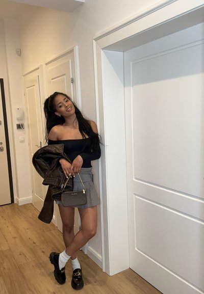 Top negro sin hombros, mini falda a cuadros grises, chaqueta de cuero marrón, bolso pequeño y mocasines negros con acentos dorados. Suelo de madera clara.