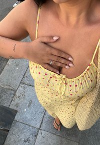 Κίτρινο φλοράλ φόρεμα με ελαστική λαιμόκοψη, μικρά ροζ και πράσινα σχέδια, και ένα υφαντό μπεζ τσάντα. Γυμνά πόδια με ανοι�χτό ροζ βερνίκι νυχιών.