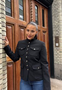 Svart skreddersydd jakke med høy krage, fire lommer foran og metallknapper. Brukt med blå jeans foran en teksturert murvegg.