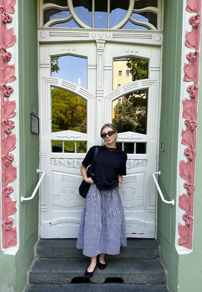 Camiseta negra, falda de longitud media con estampado de cuadros, zapatos planos negros, de pie junto a una puerta de entrada blanca ornamentada con una fachada verde y rosa.