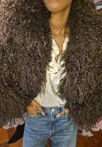 Barna göndör szőrme dzseki texturált felülettel, fehér fodros blúz fölött viselve, kék magasderekú farmerrel párosítva.