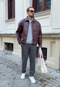 Bărbat îmbrăcat cu o jachetă de piele maro, pulover gri, pantaloni negri, teniși albi, ochelari de soare negri, ținând o geantă de pânză bej pe trotuarul din oraș.