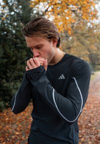 Černé atletické tričko s dlouhým rukávem, stříbrnými akcenty a perforovanou texturou. Tričko má logo na hrudi a přiléhavé rukávy.