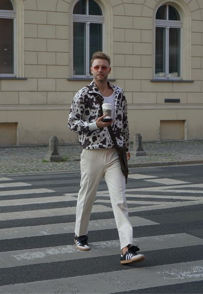 Un hombre camina por un paso de cebra, vistiendo una chaqueta de manchas en blanco y negro, pantalones blancos y zapatos Adidas negros, sosteniendo una taza de café.