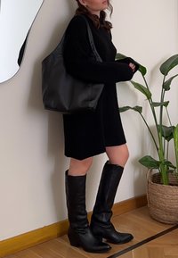 Bolsa tote de couro preto com uma textura suave, combinada com um vestido suéter preto largo e botas altas pretas, em frente a um fundo de plantas.