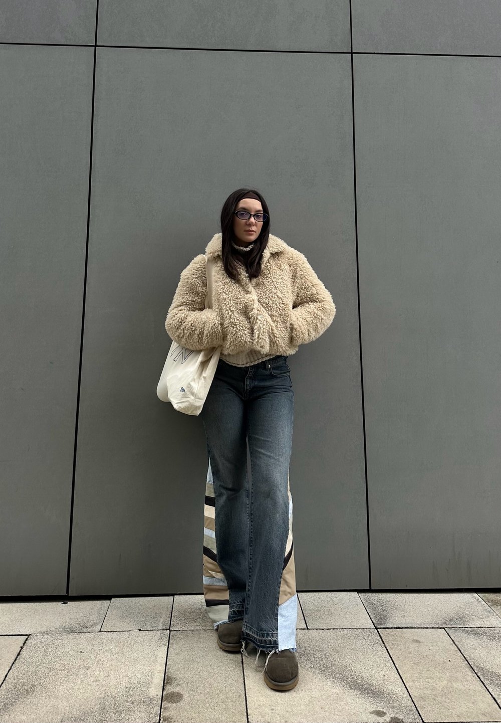 Μπεζ ψεύτικο γούνα μπουφάν, χαλαρές σκούρες τζιν με πάνελ από μπαλώματα στο πλάι, καφέ παπούτσια και μια λευκή τσάντα tote σε έναν γκρίζο τοίχο.