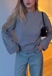 Pull en maille gris avec des poignets et un ourlet côtelés, associé à un jean bleu clair. Le tissu semble doux avec une texture confortable.