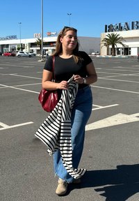 Una donna indossa una t-shirt nera e jeans blu a vita alta, tiene in mano uno scialle a righe e una borsa rossa, con scarpe beige. Sfondo di un parcheggio illuminato dal sole.