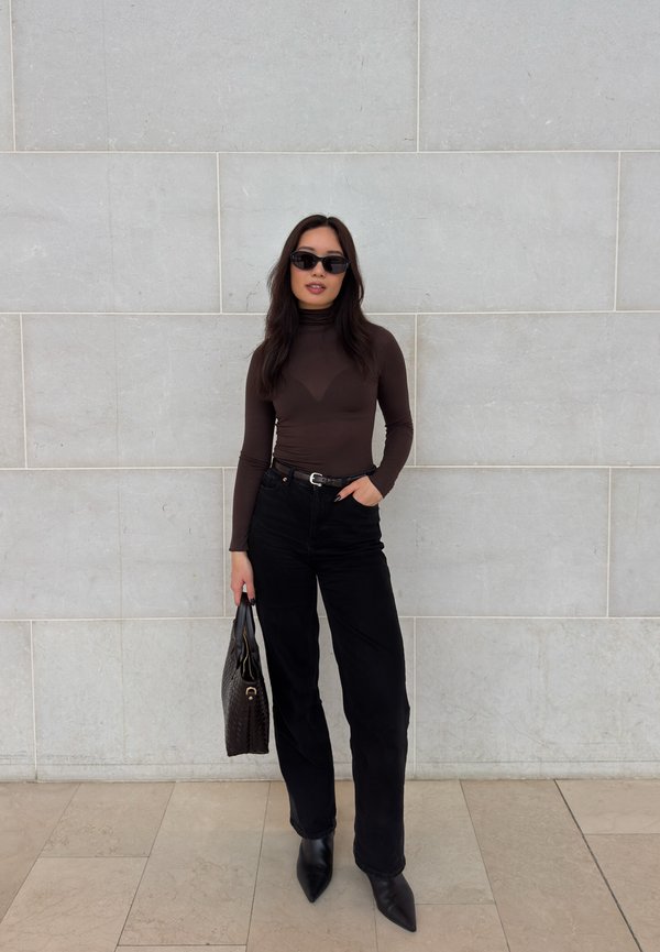 Žena v slnečných okuliaroch, oblečená v hnedom roláku a čiernych nohaviciach, držiaca čiernu kabelku, stojaca pred svetlosivou stenou s dlaždicami.