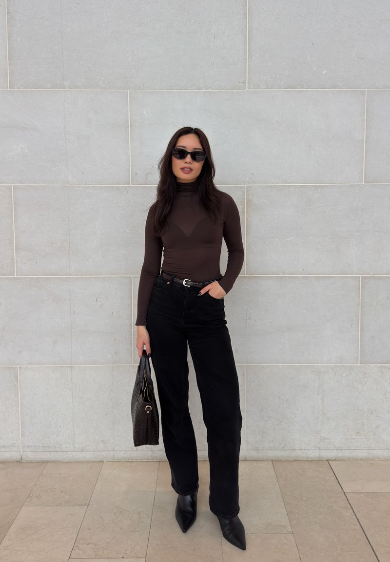 Γυναίκα με γυαλιά ηλίου, ντυμένη με καφέ ζιβάγκο και μαύρο παντελόνι, κρατώντας μια μαύρη τσάντα, στέκεται μπροστά σε ένα ανοιχτό γκρι τοιχο με πλακάκια.