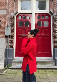 Maglione rosso a coste con colletto alto e spacchi laterali, abbinato a jeans scuri, in piedi davanti a porte doppie rosse e un muro di mattoni.