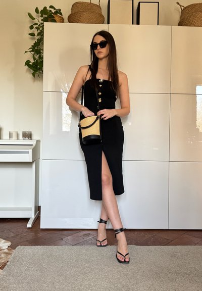 Mujer con vestido negro sin mangas, con botones y abertura, gafas de sol negras, tacones negros con tiras, sosteniendo un bolso beige y negro en interiores.