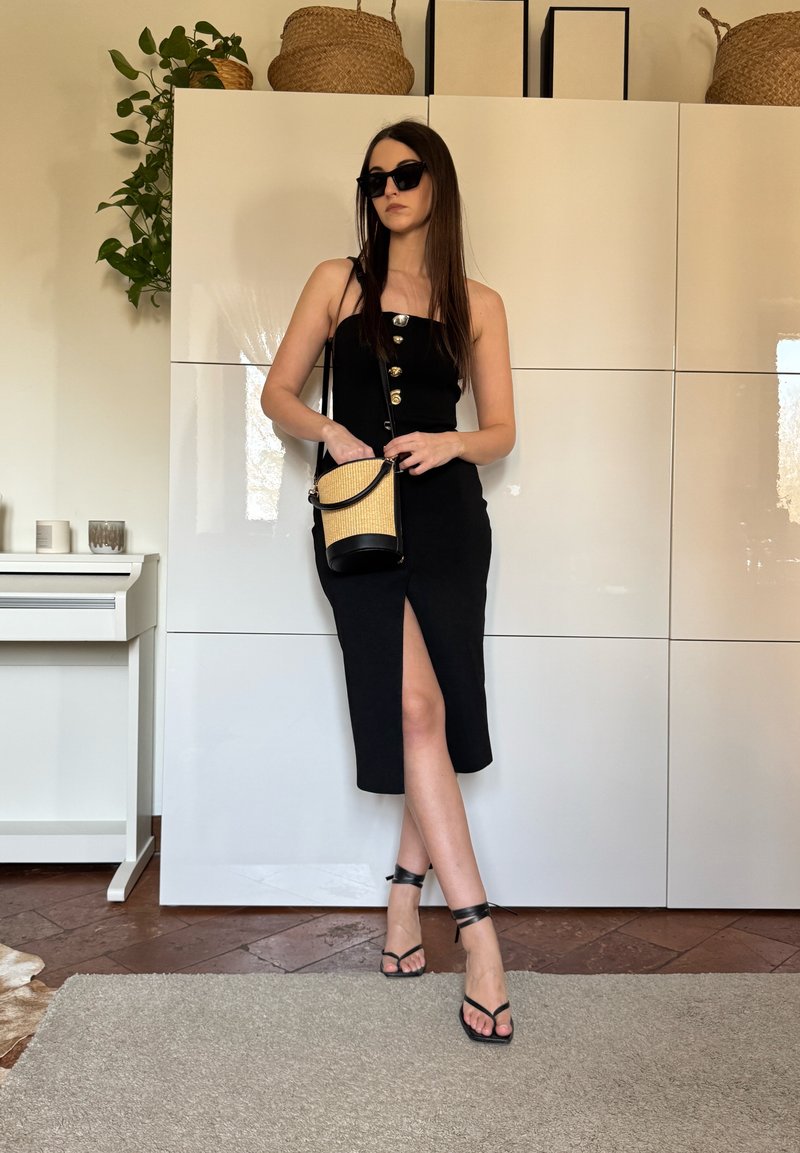 Femme portant une robe noire sans manches avec des boutons et une fente, des lunettes de soleil noires, des talons noirs à lanières, tenant un sac à main beige et noir à l'intérieur.