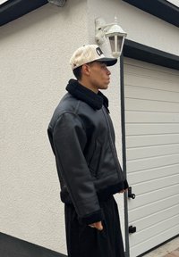 Veste en cuir noir avec un col et des poignets noirs duveteux, dotée d'une fermeture éclair à l'avant et de poches latérales, portée avec une casquette de baseball beige.