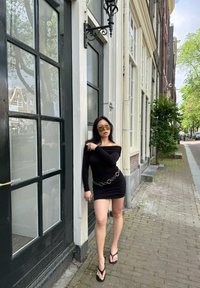 Abito nero off-shoulder con maniche lunghe e cintura a catena circolare, abbinato a ciabatte nere. Occhiali da sole riflettenti completano il look.