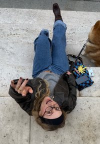 Prillidega inimene musta peapaelaga, kes kannab rohelist kampsunit ja pruuni nahkjopet, lebab heledatel kivimägede astmetel koos mitmevärvilise kotiga.