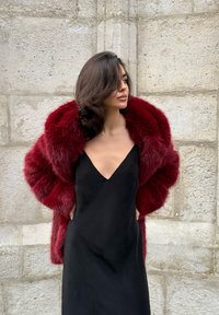 Bordeauxfarvet jakke i kunstpels med bredt sjalskrave, båret over en sort silkekjole med dybt V-udskæring. Glat tekstur med rig farve.