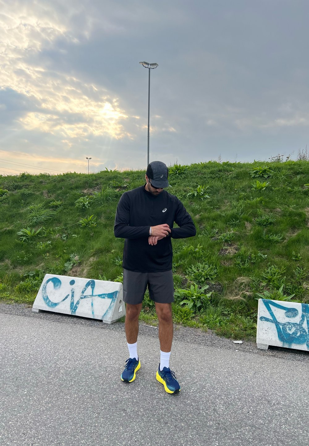 Melna garām piedurknēm krekls, pelēki šorti un zili sporta apavi ar dzelteniem akcentiem. Galvā melna cepure, un tiek pārbaudīta fitnesa pulksteņa funkcionalitāte. Fons - zāle.