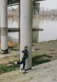 Persoon in zwarte sportkleding die één been naar achteren strekt terwijl hij bij grote betonnen pilaren naast een rivier onder een brug staat.