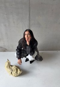 Veste en cuir noire, jupe à carreaux, pantalon blanc, bottes noires à bout pointu, en position accroupie, avec un sac vert texturé sur une surface lisse.