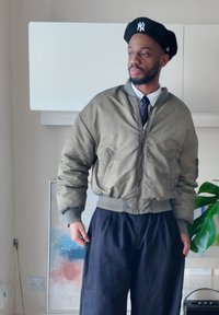 Man in olijfgroene bomberjack, zwarte broek, zwarte baret met wit logo en stropdas, staand binnen bij een plant en een abstract schilderij.