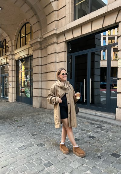 Abrigo de gran tamaño en tono marrón con grandes botones, llevado sobre una minifalda negra. Botas de plataforma marrones, gafas de sol, sosteniendo una bebida. Fondo de adoquines.