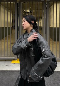 Giacca di pelle nera con tasche con zip e cinghie, caratterizzata da una vestibilità oversize. Una borsa nera è appoggiata su una spalla contro uno sfondo grigio.
