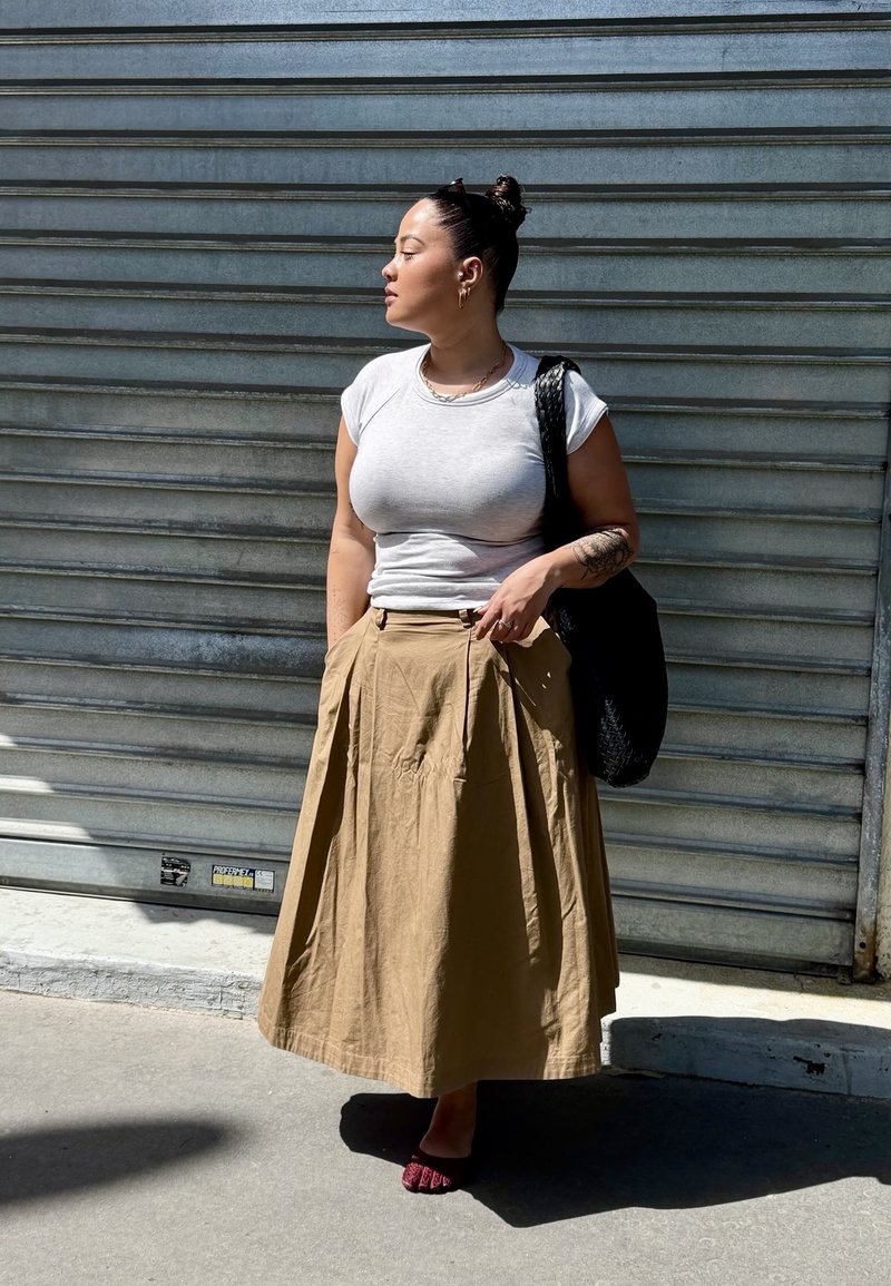 Femme avec un chignon portant un t-shirt blanc et une jupe beige, debout les mains dans les poches contre un volet métallique, portant un sac tressé noir.