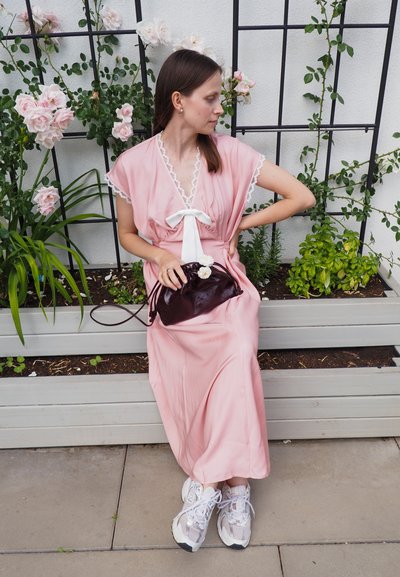 Vestido largo rosa con detalles de encaje, lazo blanco y tela fluida; bolso de mano púrpura con acentos florales; zapatillas gris; fondo de vegetación y rosas.
