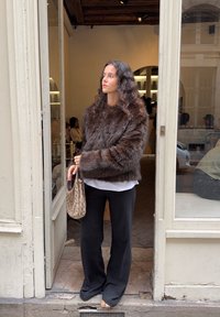 Giacca in pelliccia sintetica marrone con scollatura ampia e arrotondata, abbinata a una blouse bianca e pantaloni neri a gamba larga; tiene una borsa a fantasia.