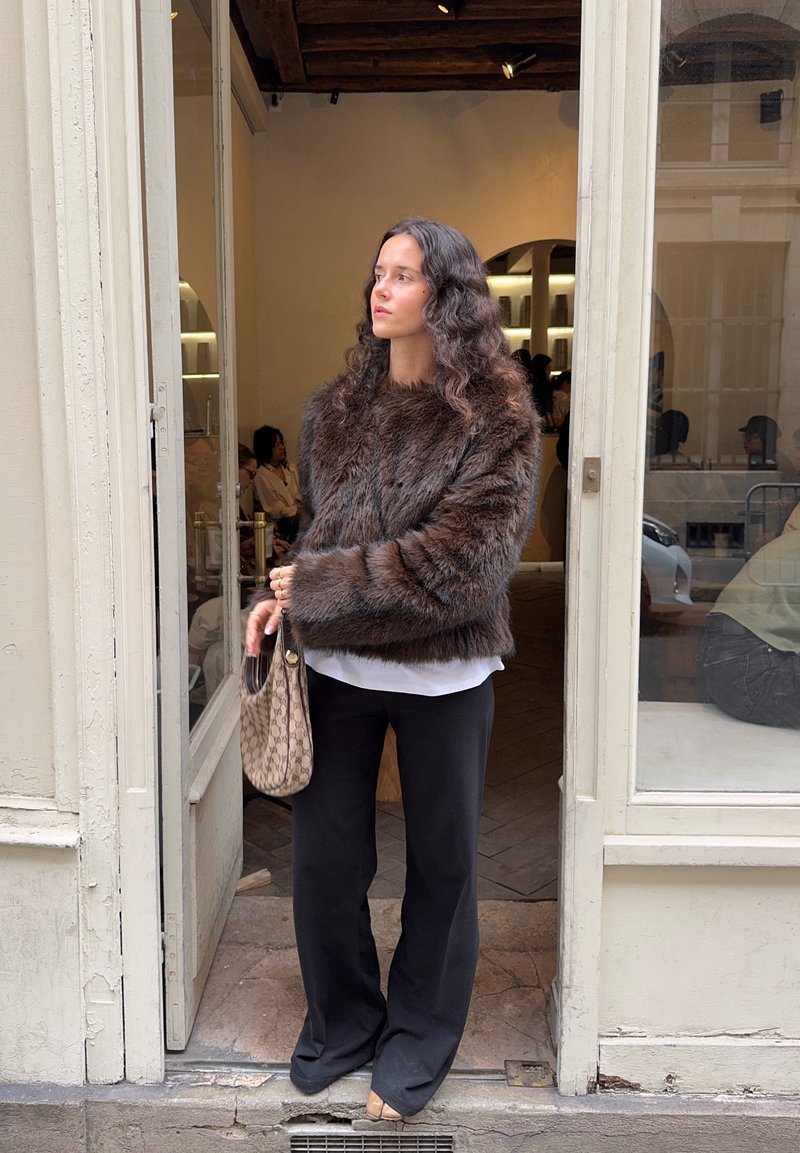 Brown faux fur jacket with a wide, rounded neckline, paired with a white blouse and black wide-leg pants; holding a patterned bag.