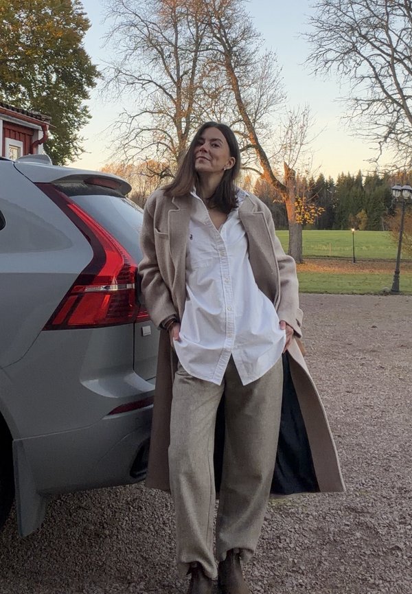 Beige ullfrakk over en hvit skjorte med knapper, paret med lysebeige bukser og brune ankelstøvler. Grå SUV i bakgrunnen.