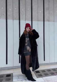 Čierny dlhý kabát z umelého kožúšku cez sivé maskáčové šaty na športový spôsob, doplnené čiernymi čižmami s strapcami a červenou pletenou čiapkou, na vzorovanom pozadí.