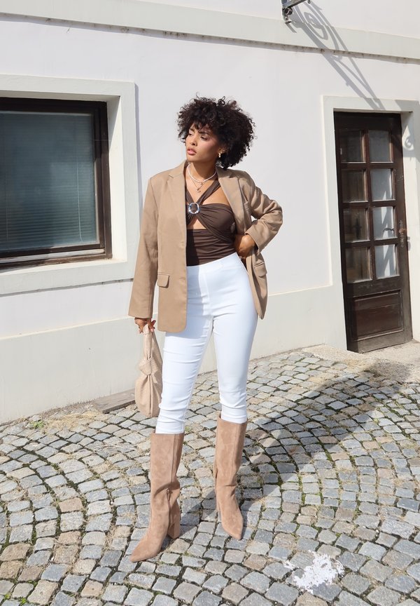 Béžový blazer přes hnědý top, bílé slim-fit kalhoty a béžové vysoké boty ke kolenům. Drží světle béžovou tašku. Stojí na dlažebních kostkách.