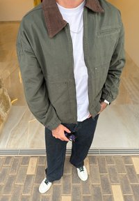 Olívazöld bombázó dzseki barna kordbársony gallérral, fehér póló, sötét farmer, fehér cipők zöld részletekkel. Napszemüveget tart a kezében.